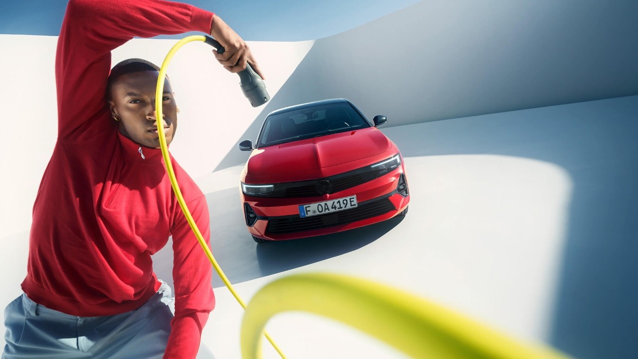 Un uomo con un cavo di ricarica EV accanto a una Nuova Opel Astra Sports Tourer Electric rossa con tettuccio nero.